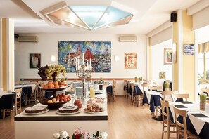 Un comedor con mesas dispuestas para una comida, una exhibición central de frutas y un cuadro grande en la pared.