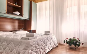 A neatly made bed with a quilted cover, a bedside table with a folded towel, a wooden shelf with decorative items, and a potted plant. - Villa Roma (Caorle)