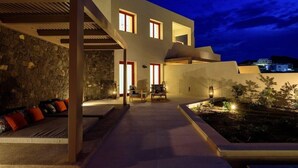 A modern house with a stone exterior, a covered outdoor seating area, and a well-lit interior. - 11:11 By Potnia Theron - Adults Only (Santorini)