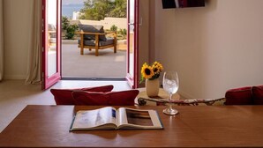 An open door leading to an outdoor seating area with a table, a book, a glass of water, and a vase with sunflowers. - 11:11 By Potnia Theron - Adults Only (Santorini)