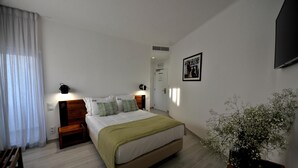 A bedroom with a bed, bedside tables, a nightstand, a window with curtains, a wall-mounted TV, and a potted plant. - Hotel Buzio (Sines)