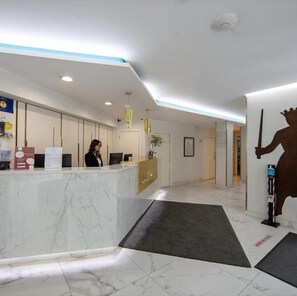 Zona de recepción de un hotel con un mostrador de mármol, una recepcionista y una escultura grande montada en la pared.