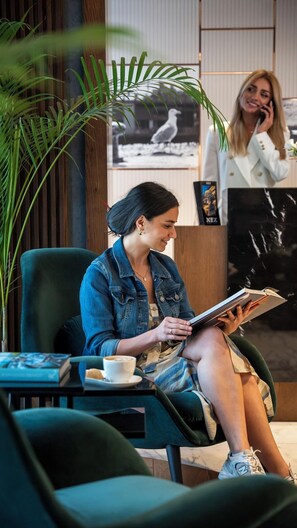 A woman sitting in a chair reading a book, with a coffee cup on a table beside her. - Nex Hotel Istanbul City Center (Istanbul)