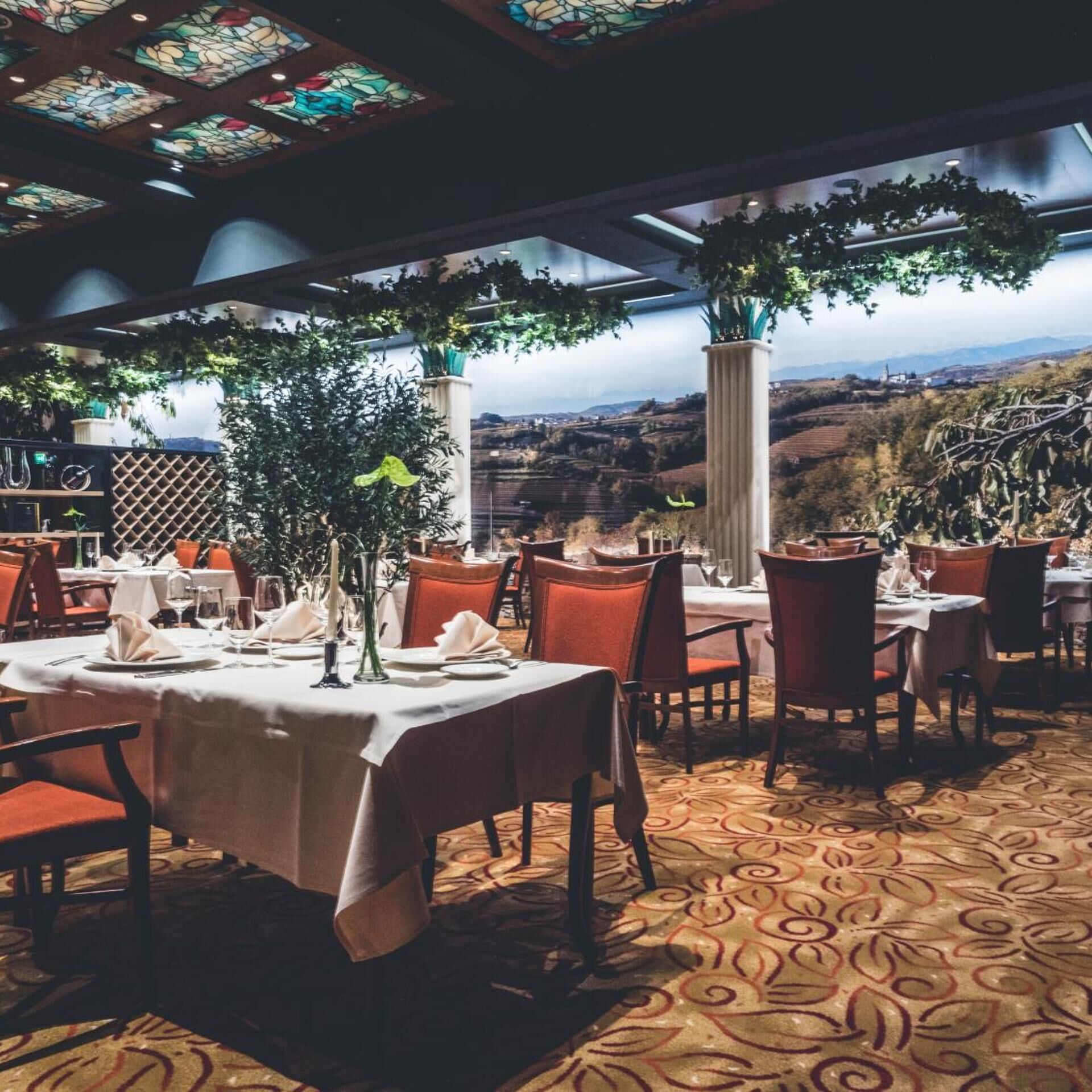 Un restaurante con mesas dispuestas para comer, vistas al campo y un techo con vidrieras de colores.