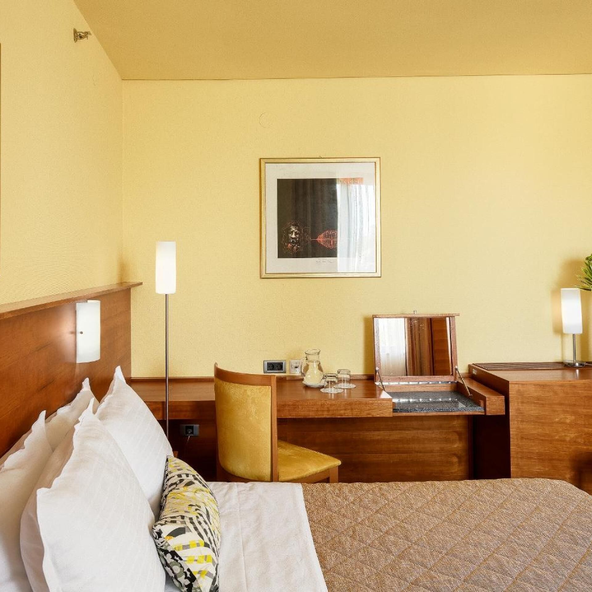A hotel room with a bed, a desk, a chair, a desk lamp, a framed picture, and a small table with a pitcher and glasses.