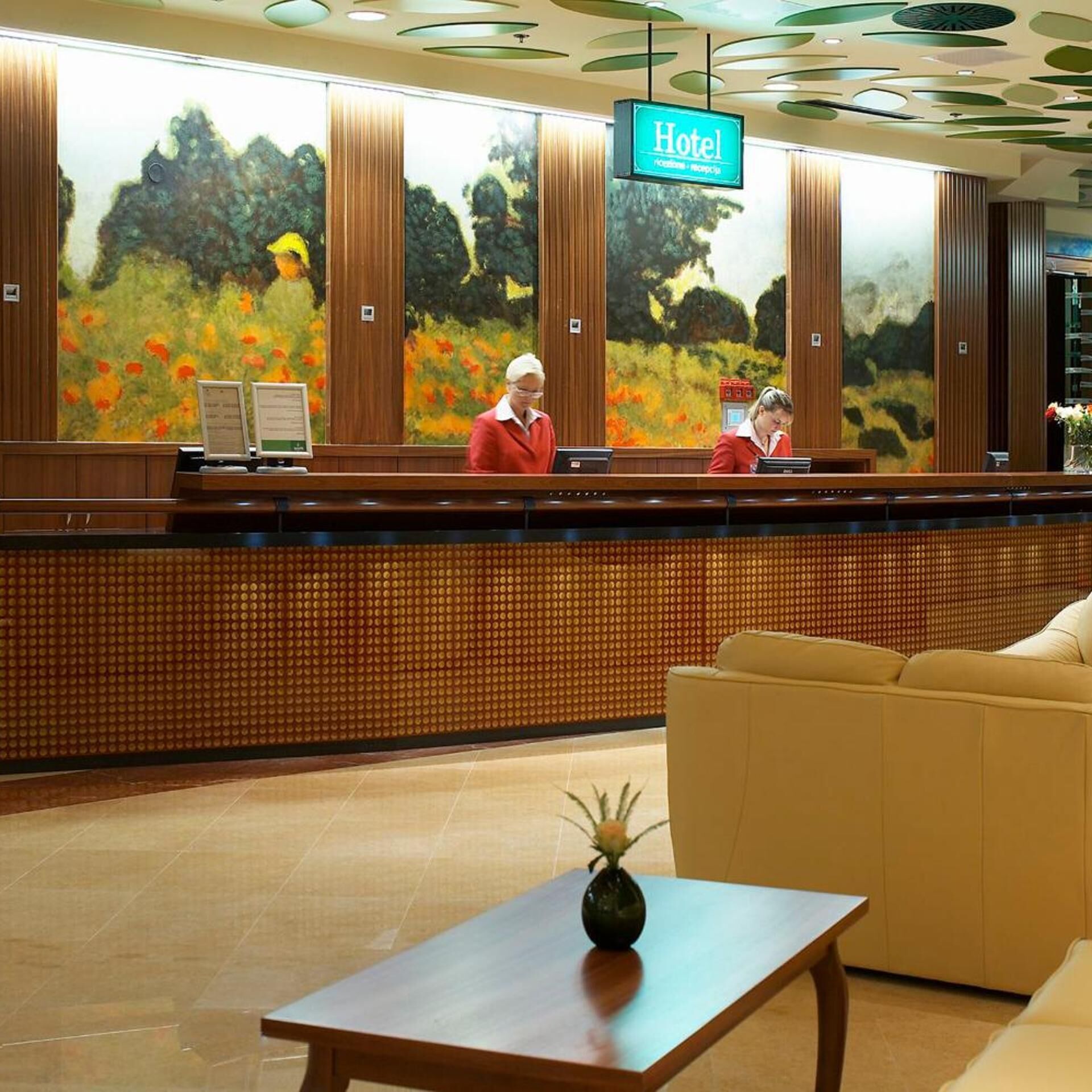 A hotel reception desk with a large painting of a landscape, two receptionists, and a reception area with a sofa and a table.