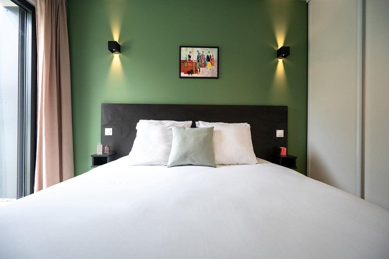 Habitación de hotel con una cama grande, paredes verdes, dos mesitas de noche con lámparas y un cuadro sobre la cama.