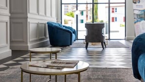 Eine Lobby mit Schachbrettfußboden, zwei runden Tischen mit Zeitungen, einem blauen Sofa und einem grauen Sessel.