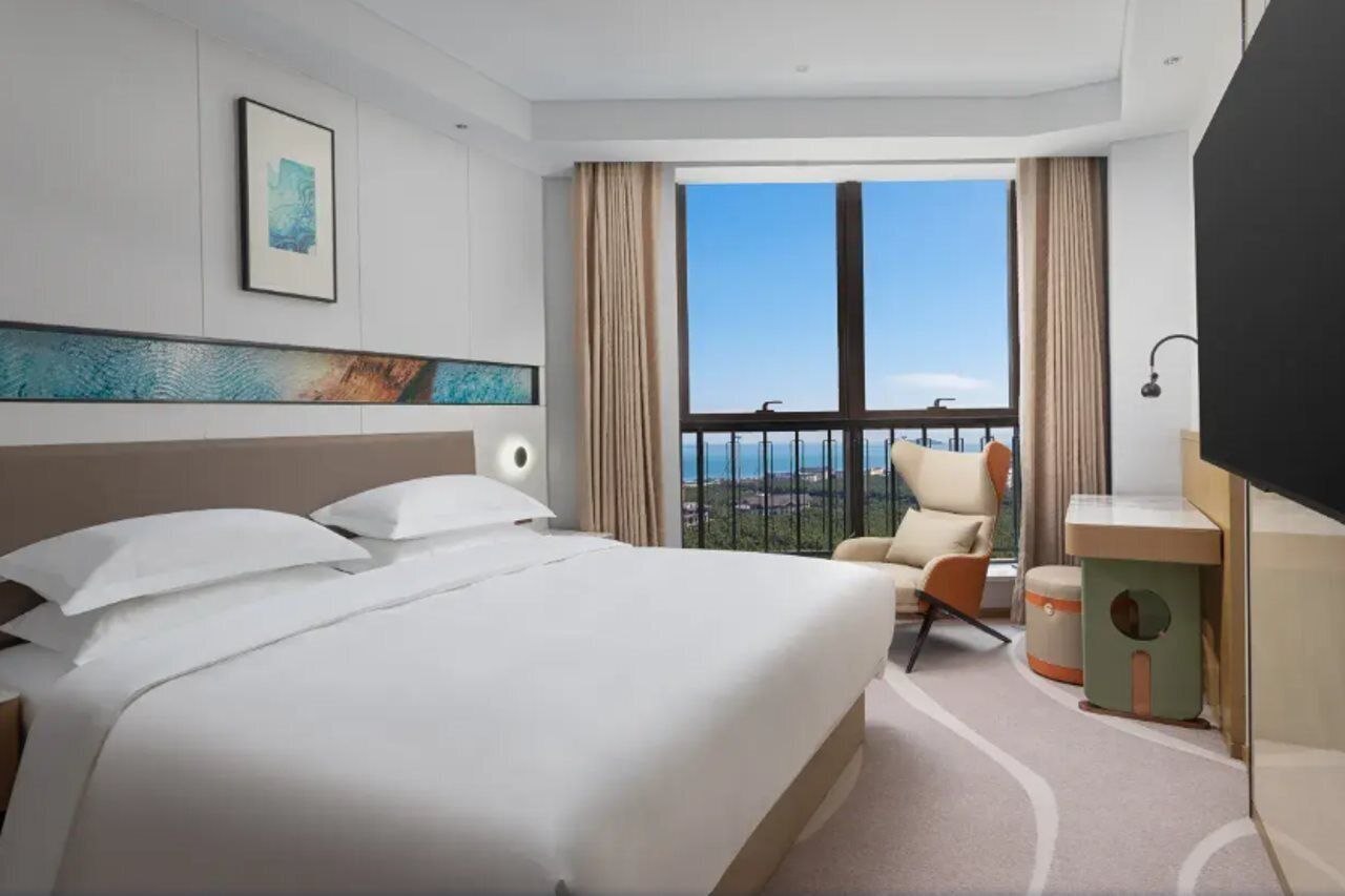 Een hotelkamer met een groot bed, een stoel, een bureau en uitzicht op de stad vanaf een balkon.