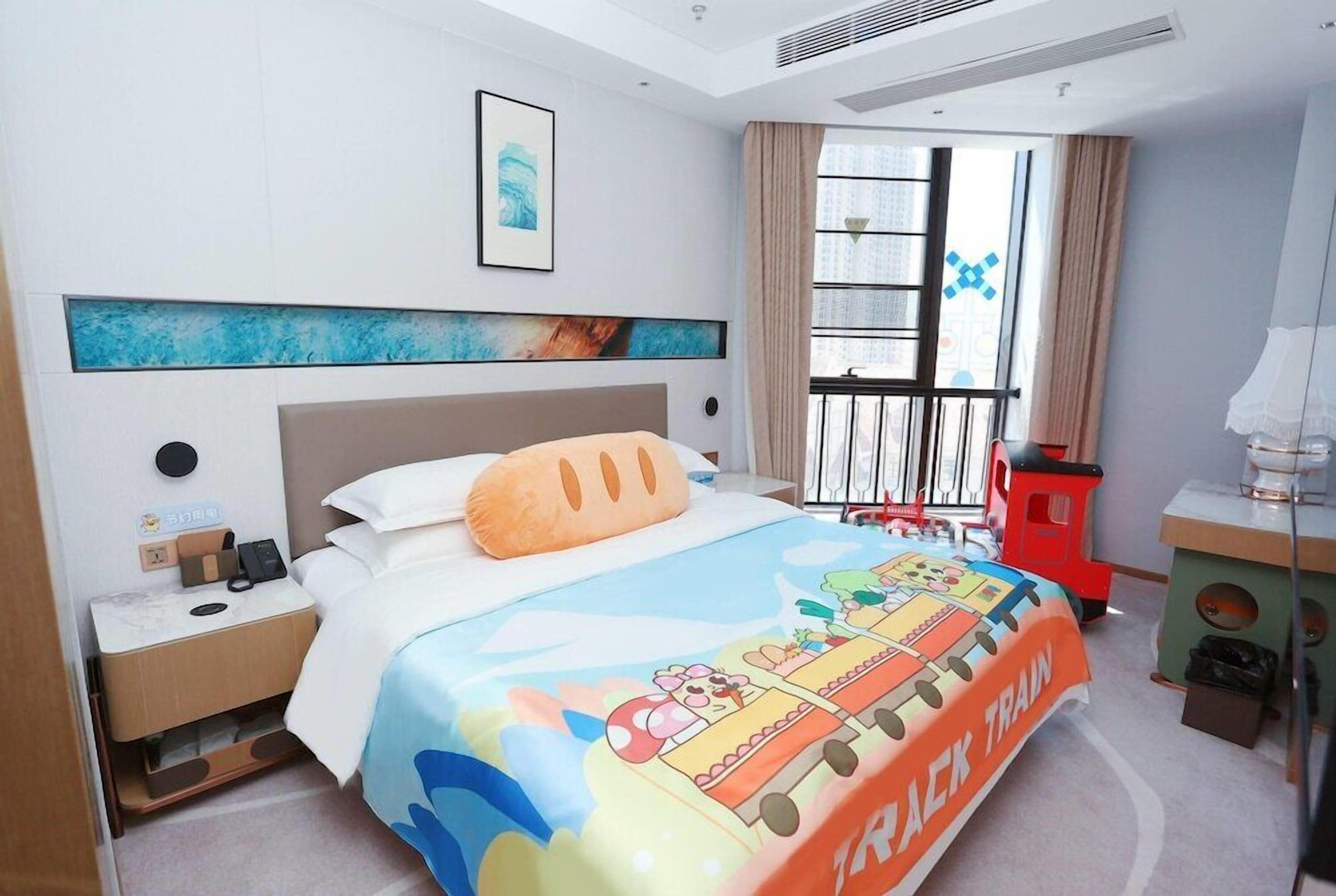 A hotel room with a bed featuring a colorful bedspread, a nightstand with a phone, a window with curtains, and a small desk with a lamp.