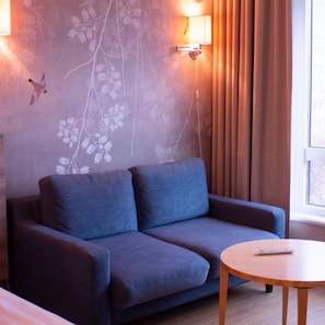 A room with a sofa, a small round table, a wall-mounted lamp, and a window with curtains. - Ulleval Hotel (Oslo)