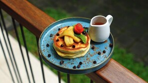 A plate with pancakes topped with fruit and chocolate chips, accompanied by a cup of syrup, on a blue plate on a wooden railing. - Inap Luxury Cabin Kintamani (Kintamani)