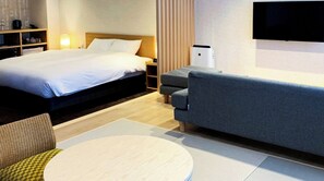 Et hotelværelse med en seng, en grå sofa, et rundt bord og et fjernsyn monteret på væggen.