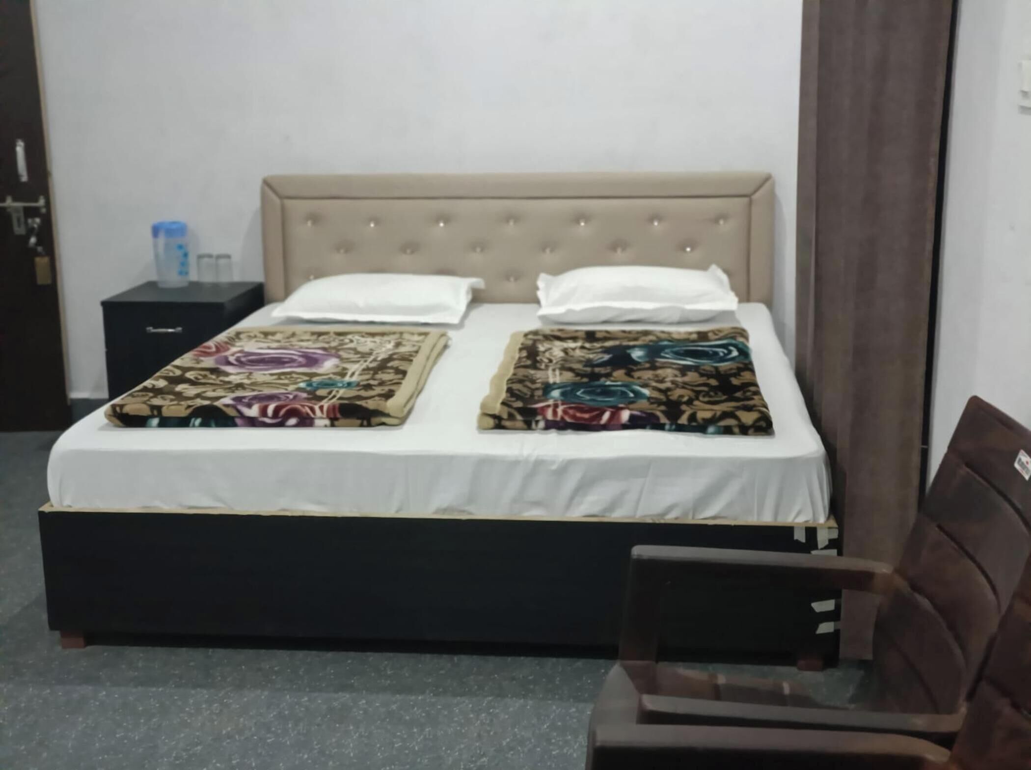 A bed with a beige headboard, two white pillows, and a floral bedspread. There is a small black nightstand with a water bottle and a tissue box. A brown chair is partially visible to the right.