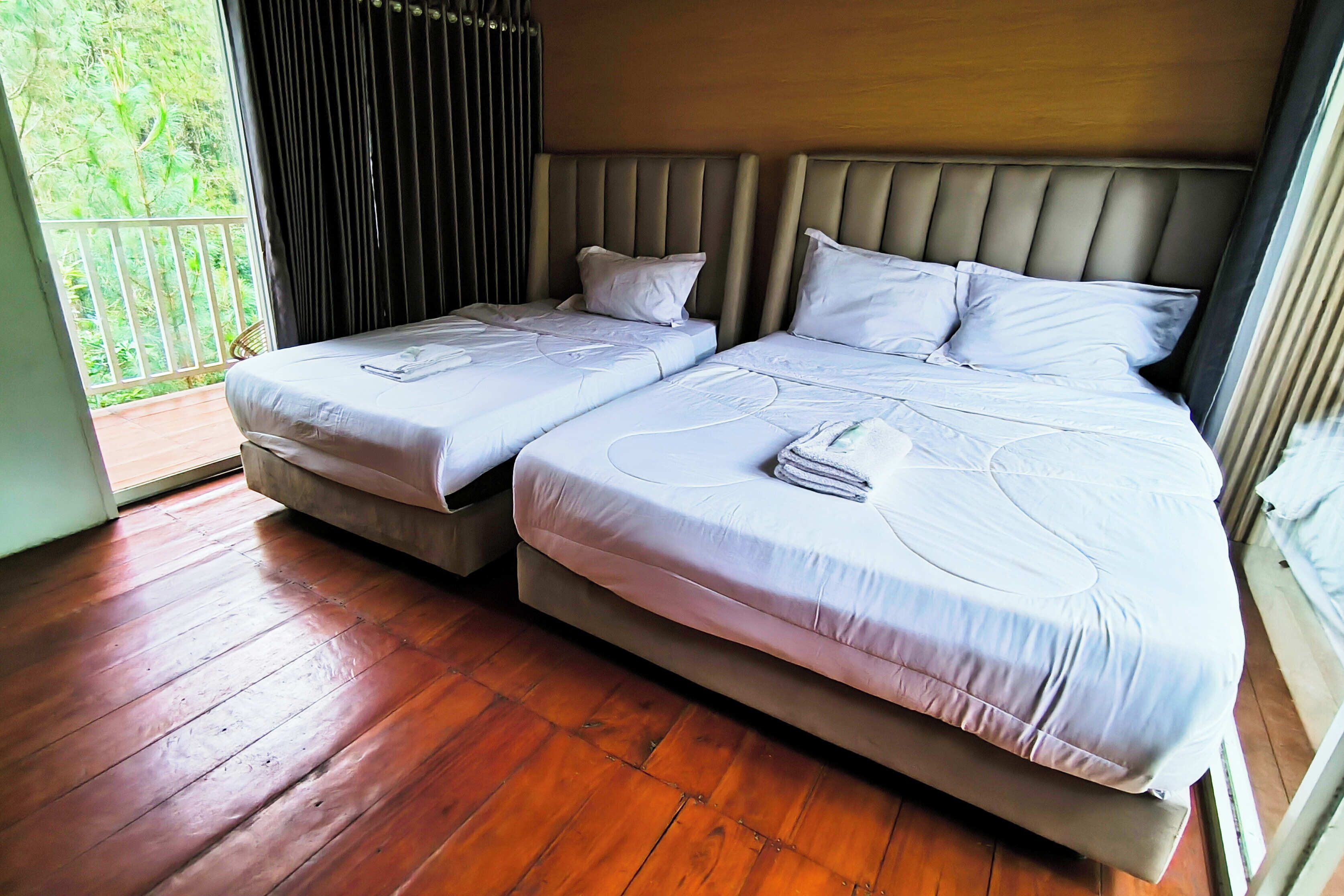 A bedroom with two beds, a wooden floor, and a view of greenery through a window.