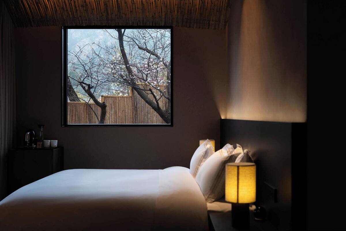 Un dormitorio con un ventanal grande, una cama, una lámpara de noche y un cuadro enmarcado en la pared.