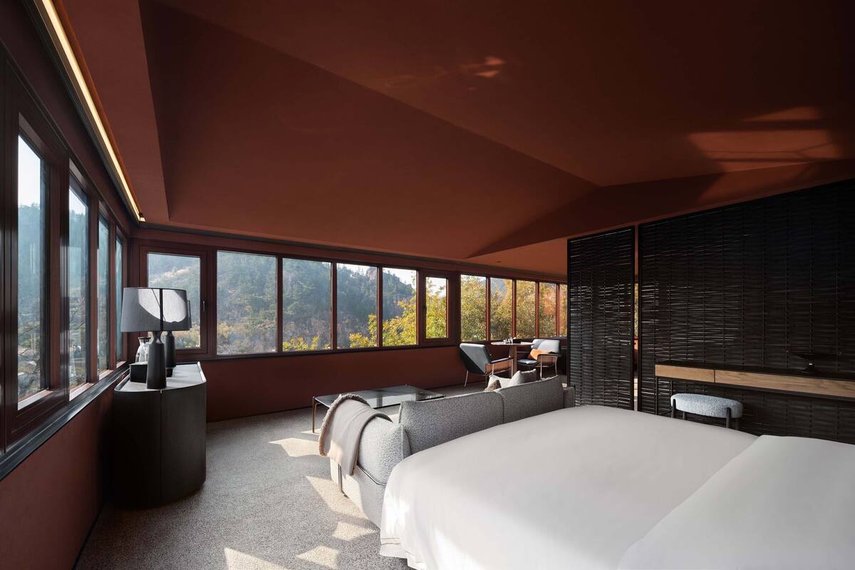 Una habitación de hotel moderna con una cama grande, una zona de estar con una mesa y vistas al exterior a través de amplias ventanas.