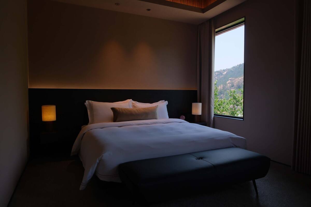 Habitación de hotel moderna con una cama grande, un banco y vistas a un paisaje montañoso por la ventana.
