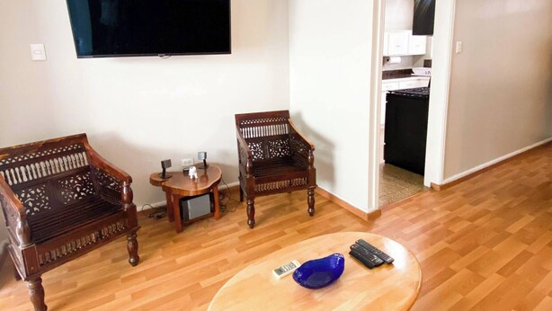 Uma sala de estar com piso de madeira, duas cadeiras de madeira, uma mesa redonda pequena com uma tigela e controle remoto, e uma TV fixada na parede.
