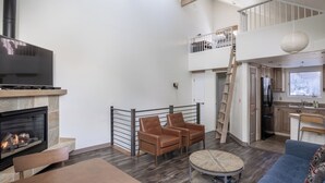 Ein großzügiger Wohnbereich mit Kamin, einem Esstisch und einem blauen Sofa. Es gibt eine Küchenzeile mit Holzschränken und einem Kühlschrank. Eine Treppe verbindet die beiden Ebenen.