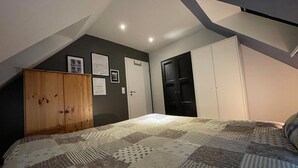 Une chambre avec un plafond en pente, une armoire en bois, un lit recouvert d’une courtepointe à motifs, une autre armoire noire et une porte dotée d’une poignée.
