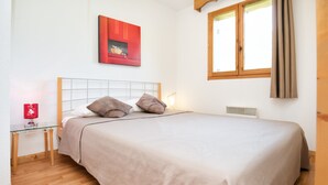 A neatly made bed with a beige bedspread and two pillows, a bedside table with a red lamp, and a window with curtains. - Apartment in 3 Vallees With Pool & Sauna (Orelle)
