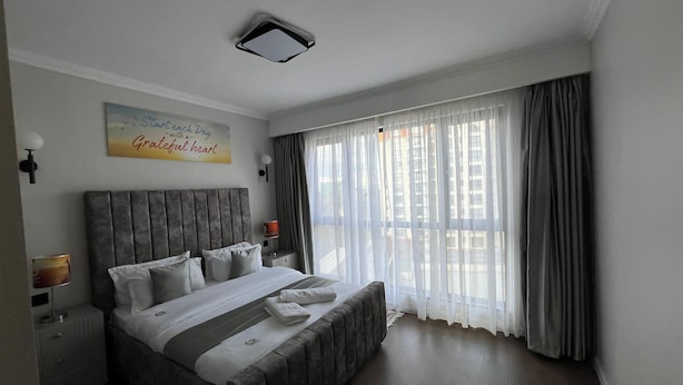 A modern hotel room with a large bed, a bedside table, a nightstand, a wall-mounted light, and a window with curtains. - Parkview Cribs. Serene 3BR with a DSQ near JKIA and SGR with National park view. (Nairobi)