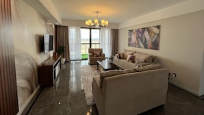 A modern living room with a sofa, coffee table, and TV. There is a large window with curtains, a chandelier, and a painting on the wall. - Parkview Cribs. Serene 3BR with a DSQ near JKIA and SGR with National park view. (Nairobi)