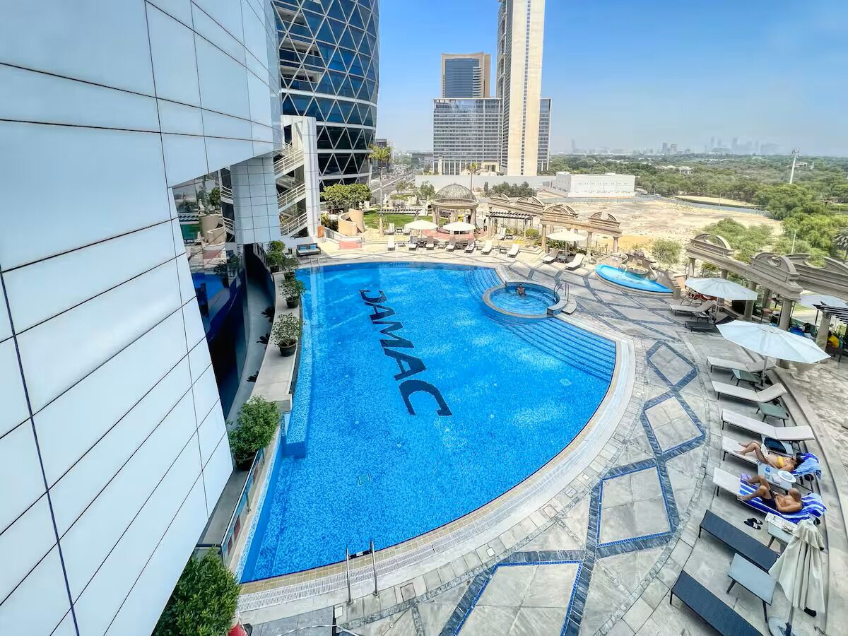 Una piscina en la azotea con la palabra "JAMAC" en el agua, rodeada de sillas de descanso y sombrillas, con un paisaje urbano de fondo.