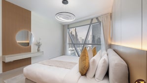 A modern bedroom with a large bed, a bedside mirror, a vase with flowers, and a view of a cityscape through a large window. - Vogue Park Tower DIFC  (Dubai)