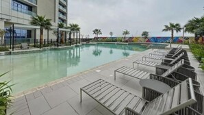 Zona de piscina de hotel con una piscina grande, sillas de descanso y un área de estar cubierta.