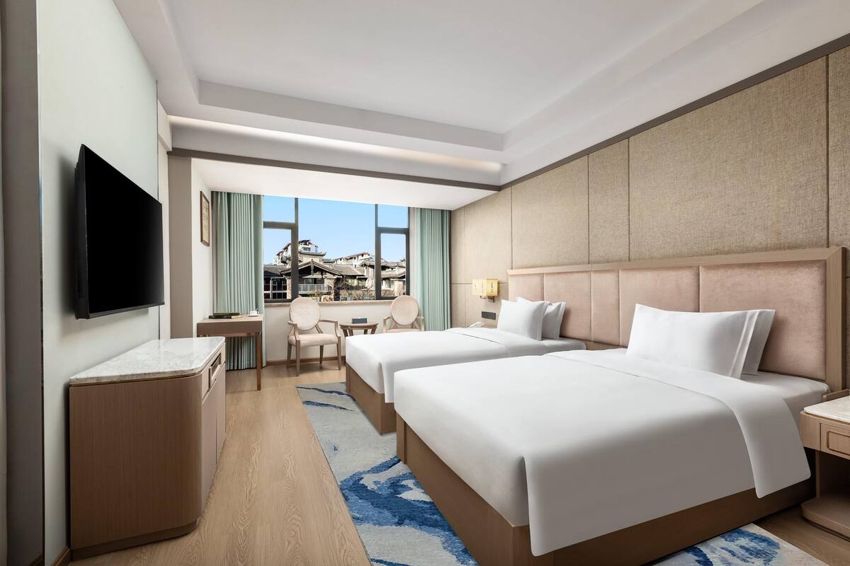 Habitación de hotel con dos camas, televisión, escritorio y vistas a edificios.