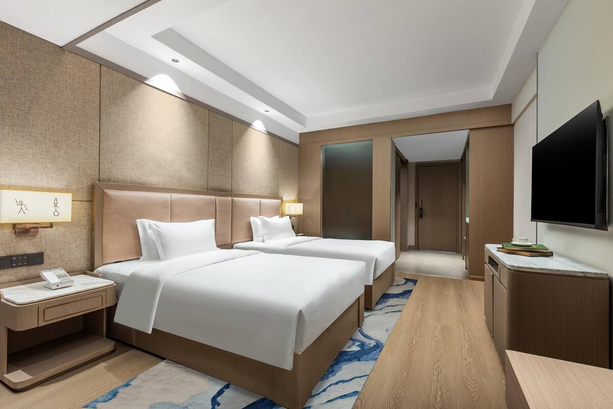 Habitación de hotel con dos camas, un escritorio, un televisor y un baño visible a través de una puerta abierta.