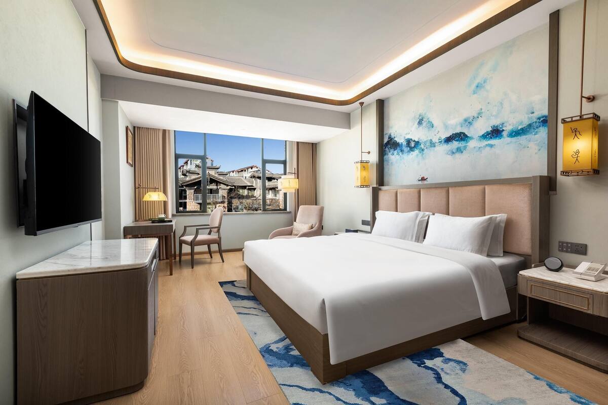 Habitación de hotel moderna con una cama grande, un escritorio, un televisor y vistas a edificios tradicionales.