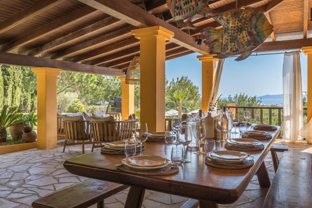 An outdoor dining area with a wooden table set for a meal, surrounded by chairs and a view of the landscape.