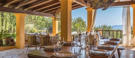An outdoor dining area with a wooden table set for a meal, surrounded by chairs and a view of the landscape.