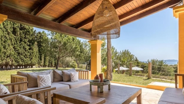 An outdoor seating area with wooden furniture and a wicker lamp, overlooking a landscaped garden and a clear view of the sea. - Villa Casa FOUR (Can Pep Simó)