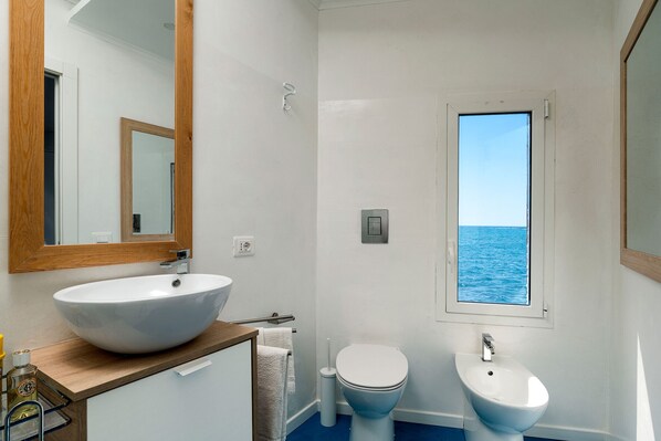Ein Badezimmer mit einem großen weißen Waschbecken, einer Toilette und einem Bidet. Ein Fenster bietet Meerblick.