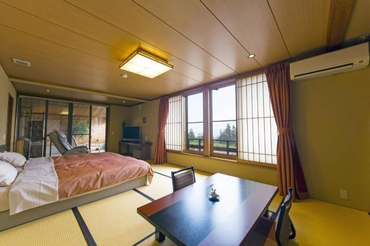 A hotel room with a bed, a dining table, a chair, a TV, and large windows with curtains.