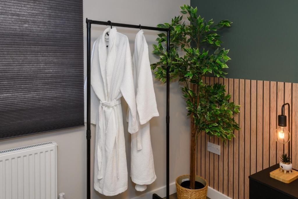 A bathroom with a towel rack holding two white bathrobes, a potted plant, a radiator, and a wall-mounted light fixture.