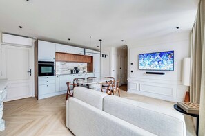 Ein modernes Wohnzimmer mit einer Couch, einem Esstisch, Stühlen, einer Küche mit weißen Schränken und einem an der Wand befestigten Flachbildfernseher.