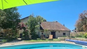 Un área de piscina con un cielo azul despejado, un edificio de piedra con techo de tejas, y una sombrilla verde.