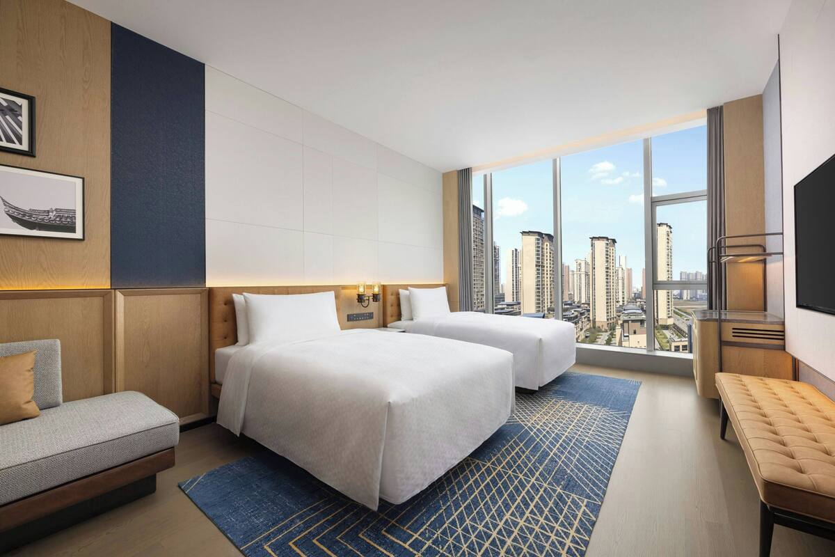 A hotel room with two beds, a sofa, a bench, a TV, and a large window with a city view.