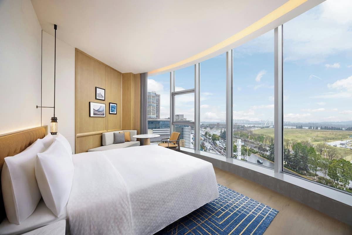 A hotel room with a large bed, a small table, a chair, and a view of a cityscape through large windows.