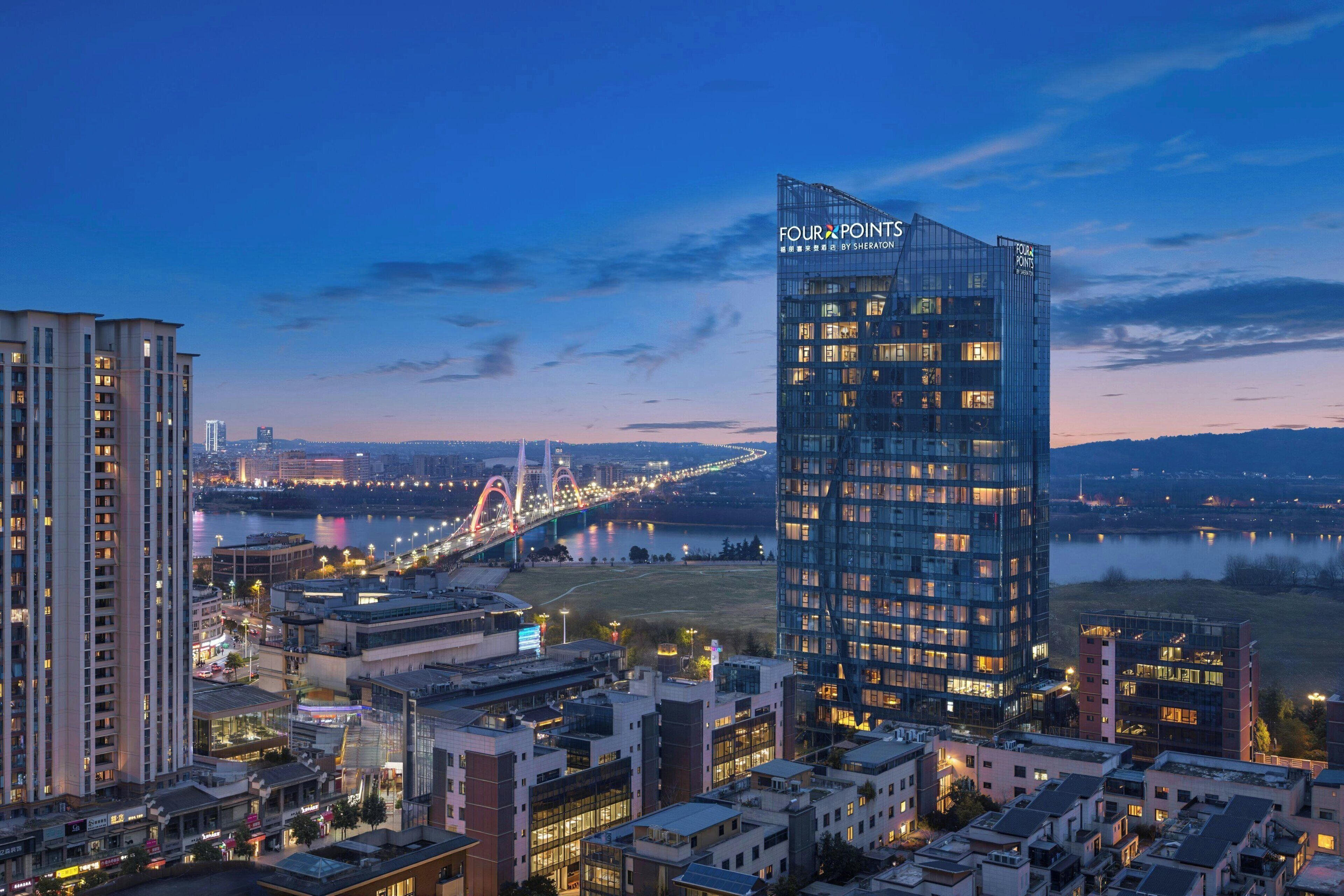 A high-rise hotel with a modern glass facade, situated in an urban setting with a river and a bridge in the background.