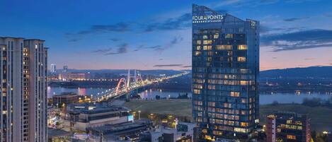 A high-rise hotel with a modern glass facade, situated in an urban setting with a river and a bridge in the background.