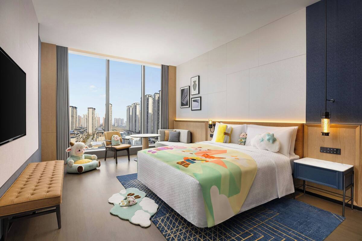 A hotel room with a large bed, a seating area with a chair and a sofa, a flat-screen TV, and a city view.