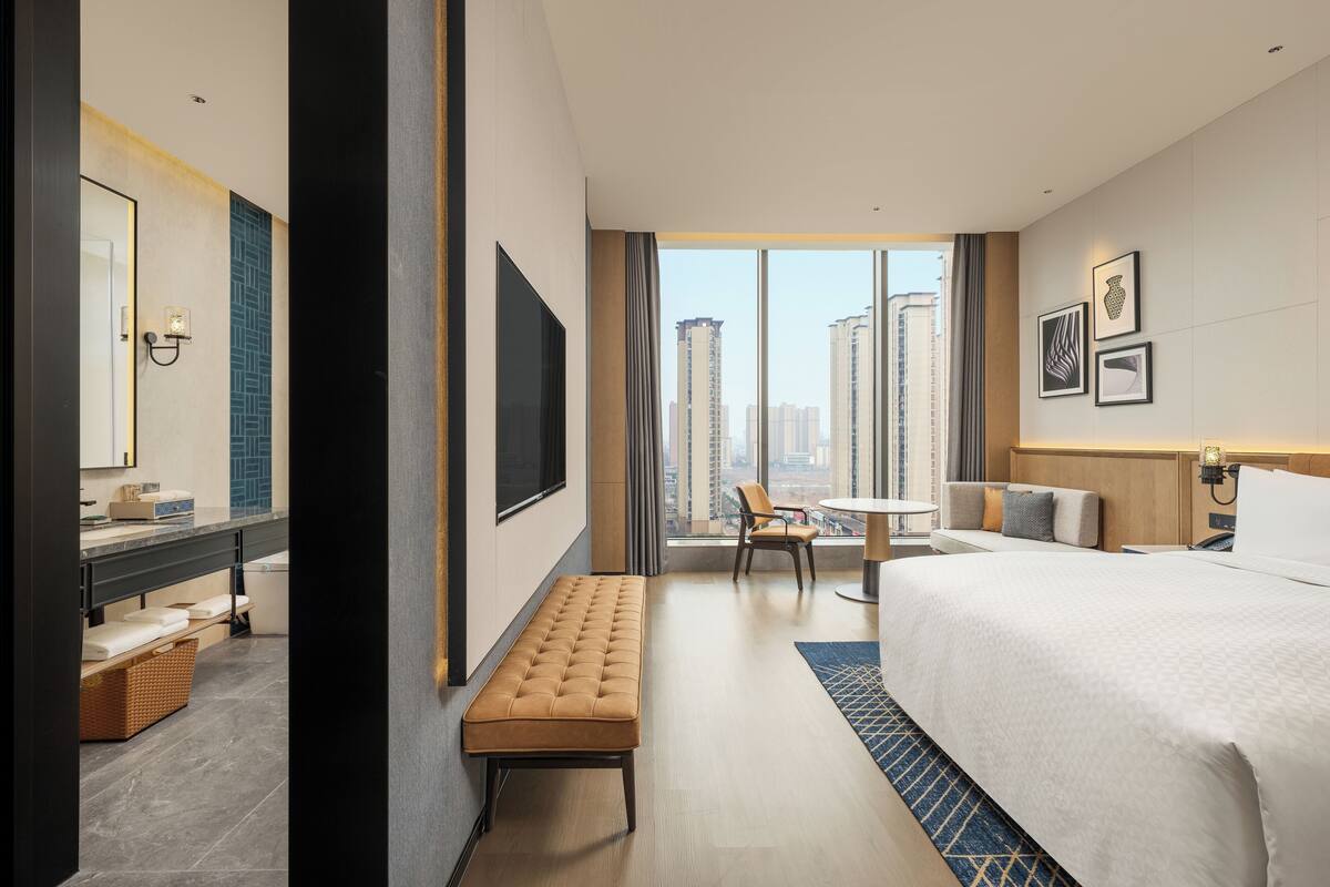 A modern hotel room with a large bed, a bench, a small table, and a view of the city.