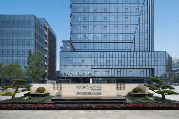 Een modern hotel met een glazen gevel, een aangelegde tuin en een bord met de tekst 'FOUR POINTS BY SHERATON'.