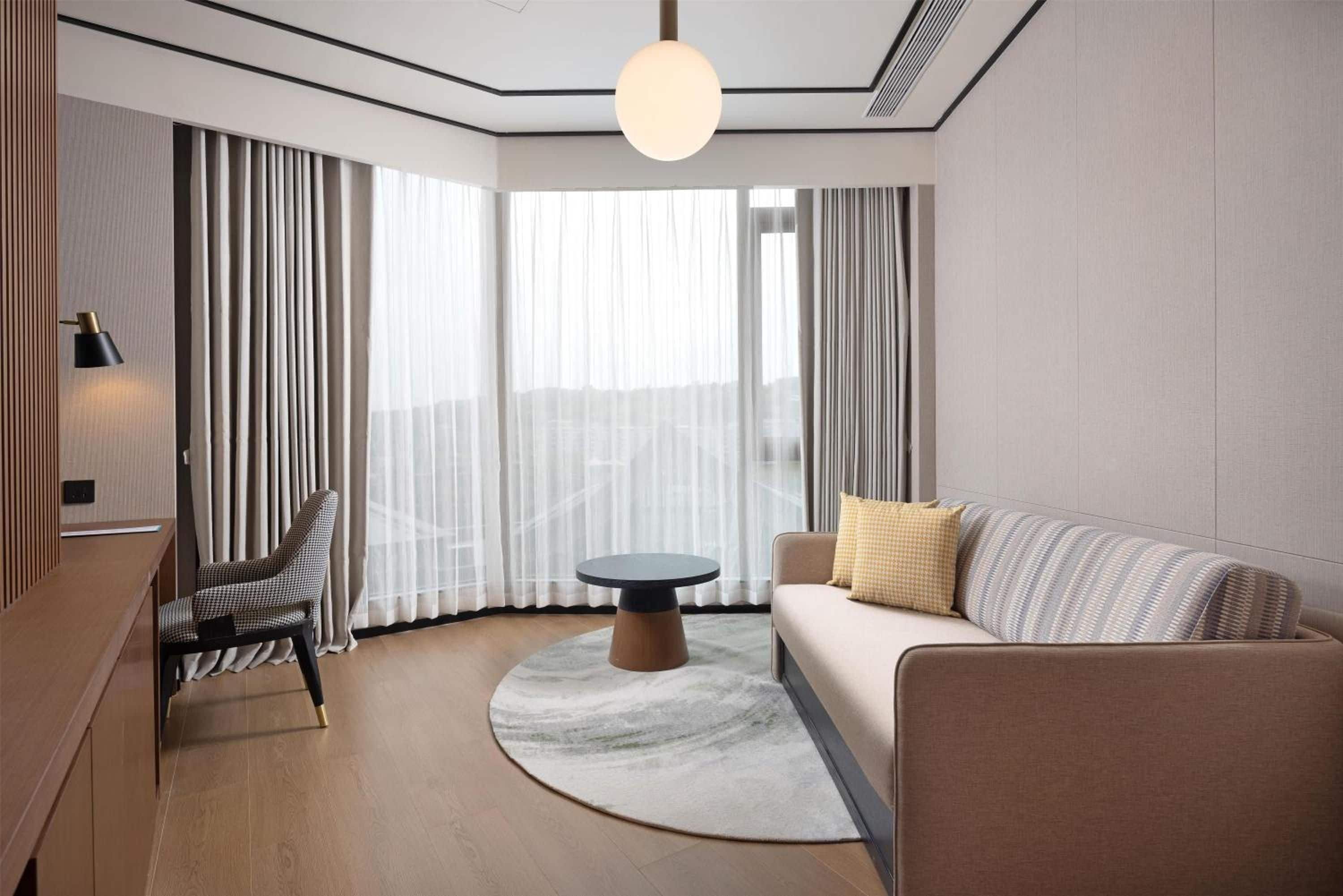 Et moderne hotelværelse med et rundt bord, en sofa, en lænestol og et stort vindue med gardiner.